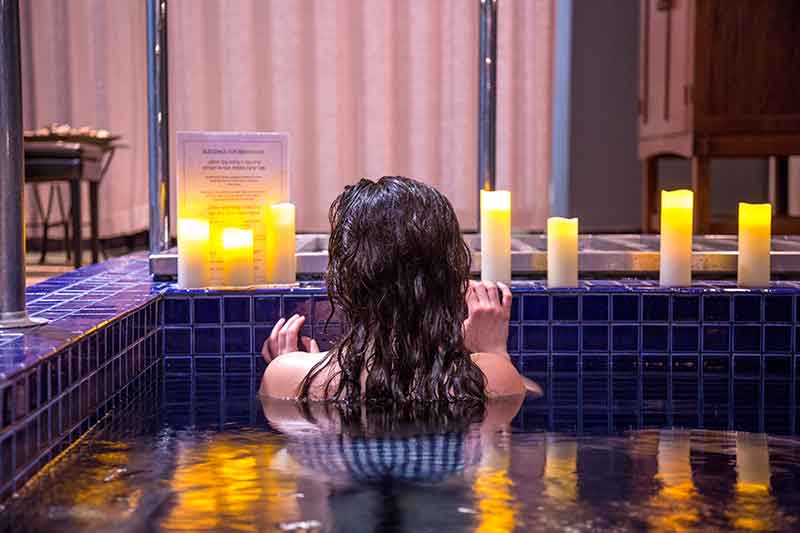 A person is soaking in a hot tub, surrounded by lit candles, with a sign on tiled edge, suggesting a tranquil, luxurious spa setting.