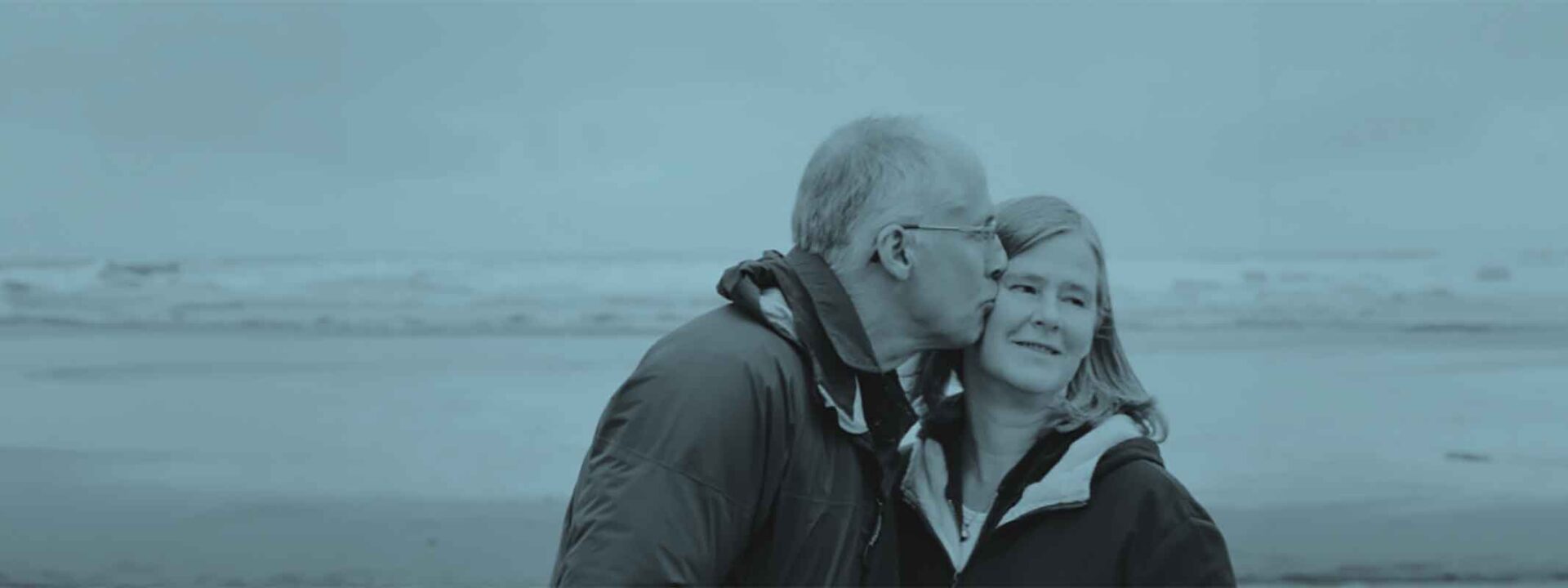 The image displays a couple embracing with one person kissing the other's forehead. They are by the sea with a vast, overcast sky.