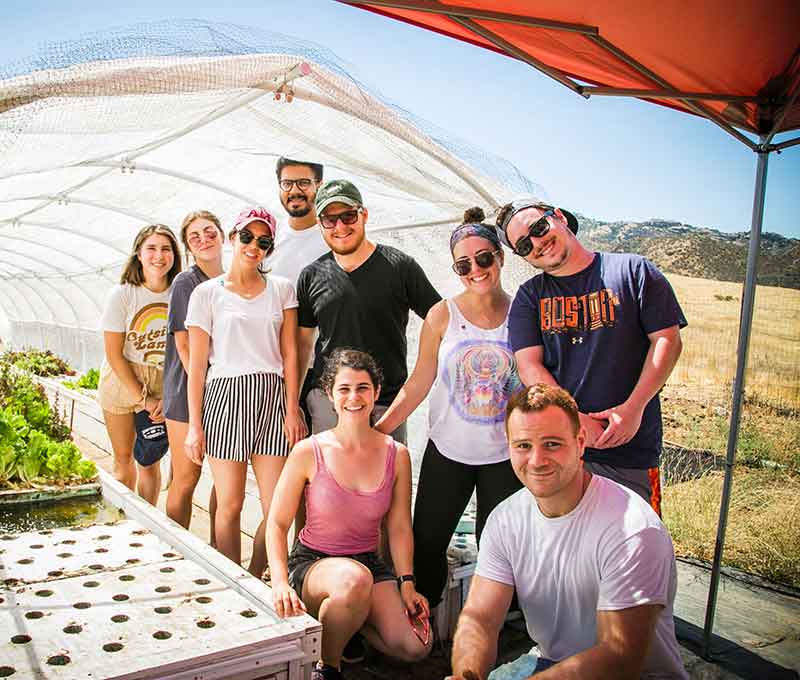 A group of nine people posing under a sunny sky with smiles, in front of agricultural equipment, with a greenhouse and a hill in the background.