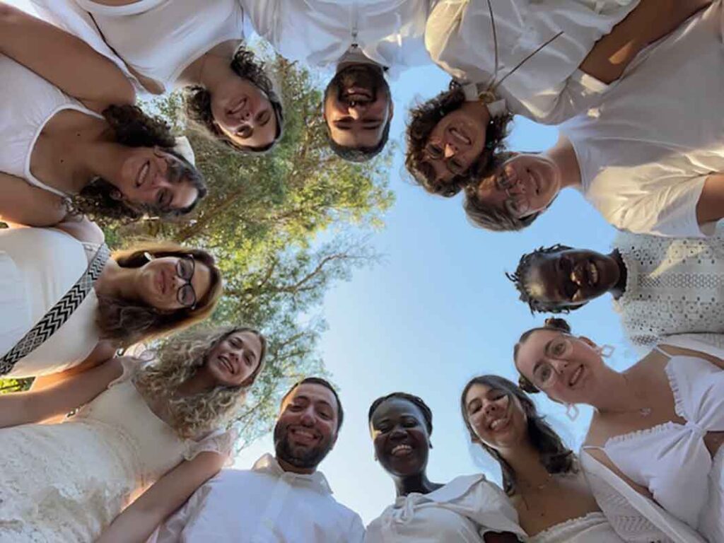 A group of individuals in white attire huddled together, smiling at the camera from a low angle, surrounded by trees under a clear sky.