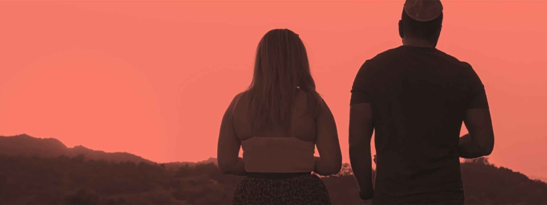 Two individuals are standing side by side against a sunset sky, overlooking a hilly landscape. The image captures a serene, warm atmosphere.