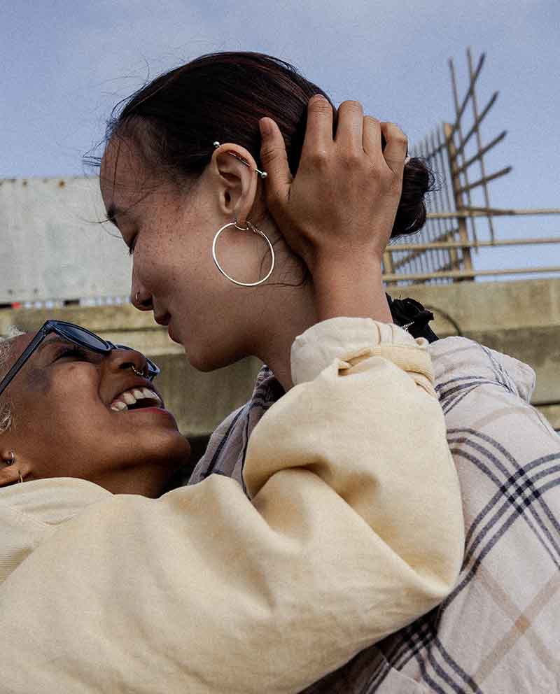The image shows two individuals close up, embracing and smiling affectionately. They are outside, with a clear sky and what appears to be construction scaffolding in the background.