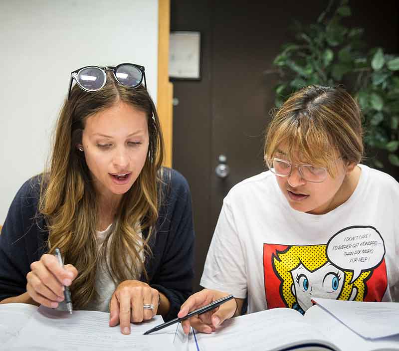 Two individuals are engaged in a discussion over documents. One is wearing sunglasses on their head; the other has a graphic T-shirt. They appear focused.