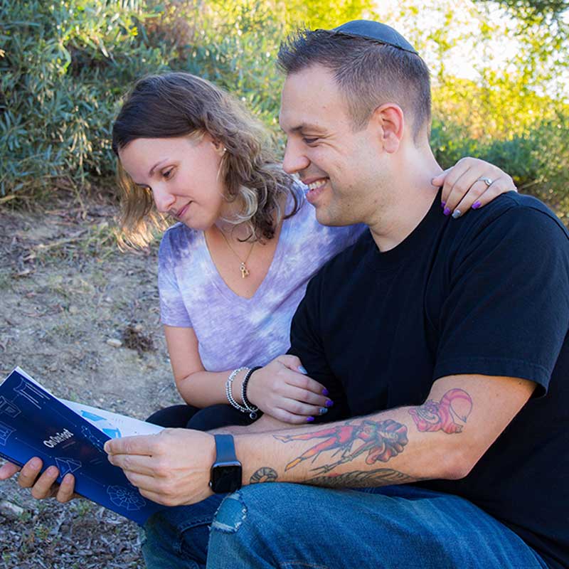 Two people are outdoors sitting closely, sharing a moment reading a book together. The person on the right has visible tattoos on their arm.
