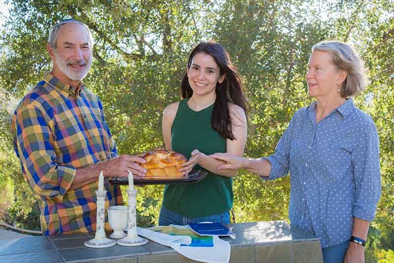 Three people outdoors with food, trees in the background. One person is holding a baked dish, a table has candles, napkins, and a beverage.