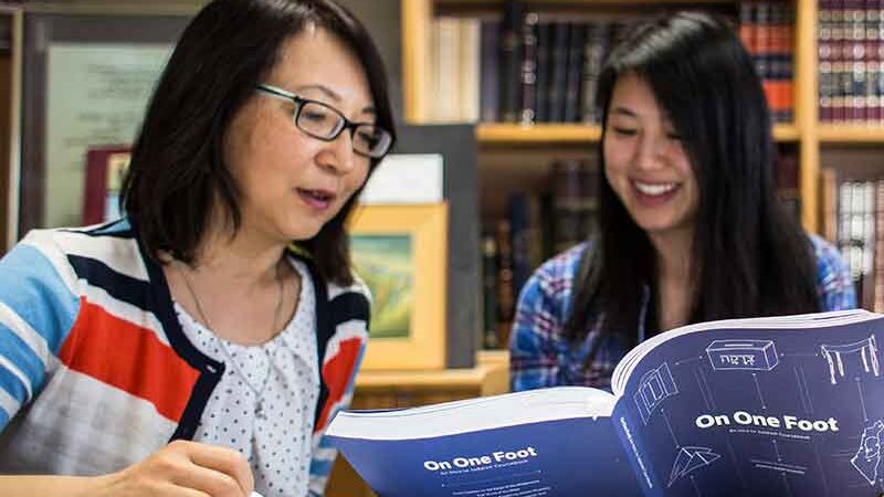 Two individuals are smiling and reading a book titled "On One Foot" in a library with bookshelves and books in the background.