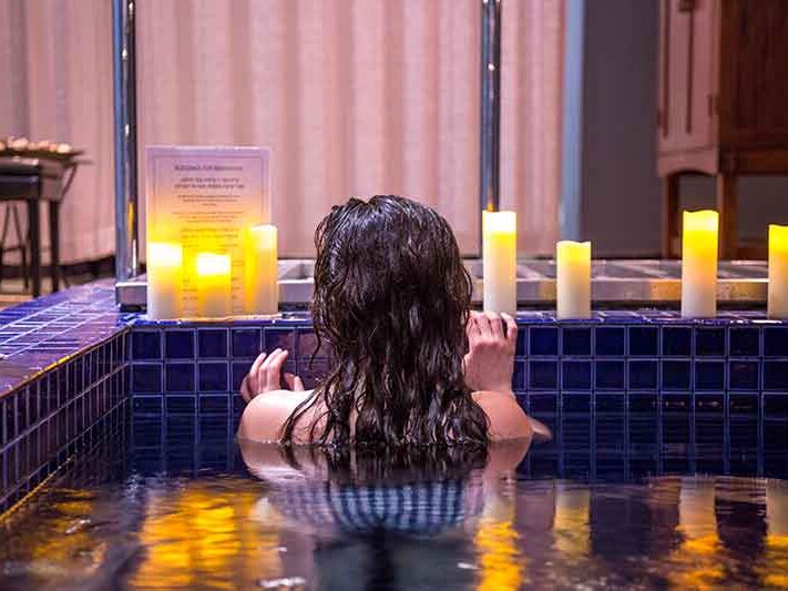 A person is in a pool surrounded by lit candles with a relaxed posture, reflecting a serene ambiance in an indoor setting with elegant decor.