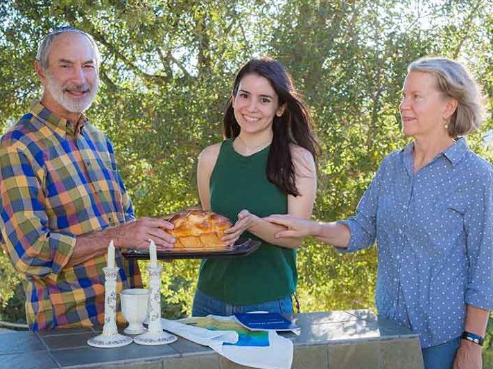 Three people outdoors with food, trees in the background. One person is holding a baked dish, a table has candles, napkins, and a beverage.