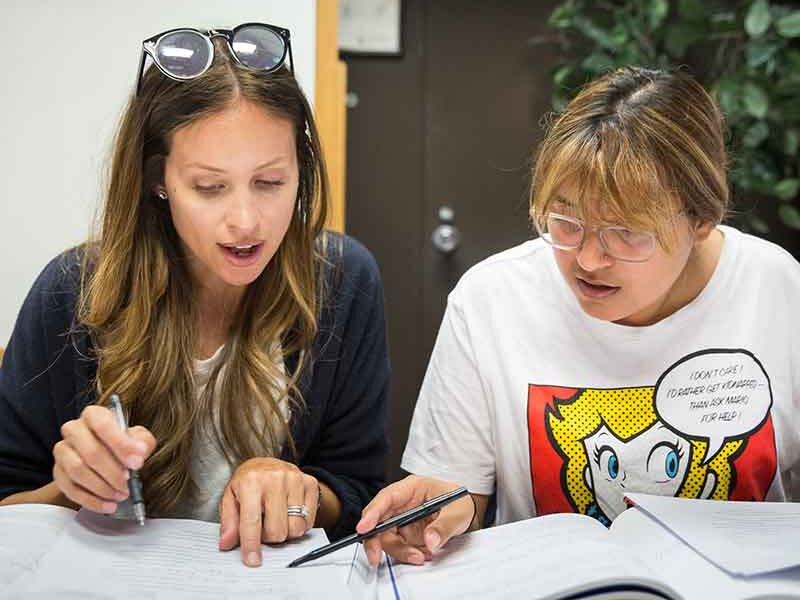 Two individuals are engaged in a discussion over documents. One is wearing sunglasses on their head; the other has a graphic T-shirt. They appear focused.